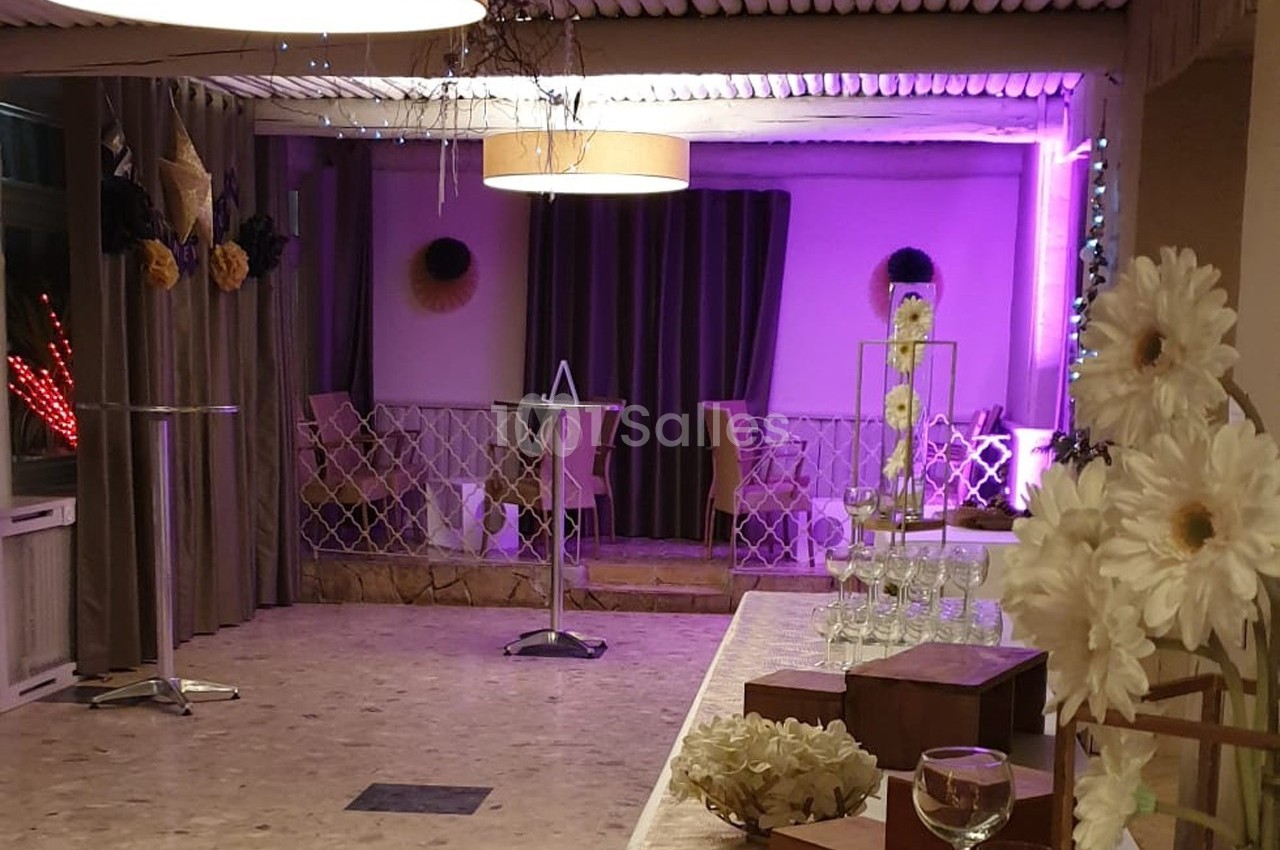 Salle décorée avec des fleurs blanches, tables hautes et éclairage violet, prête pour un événement.