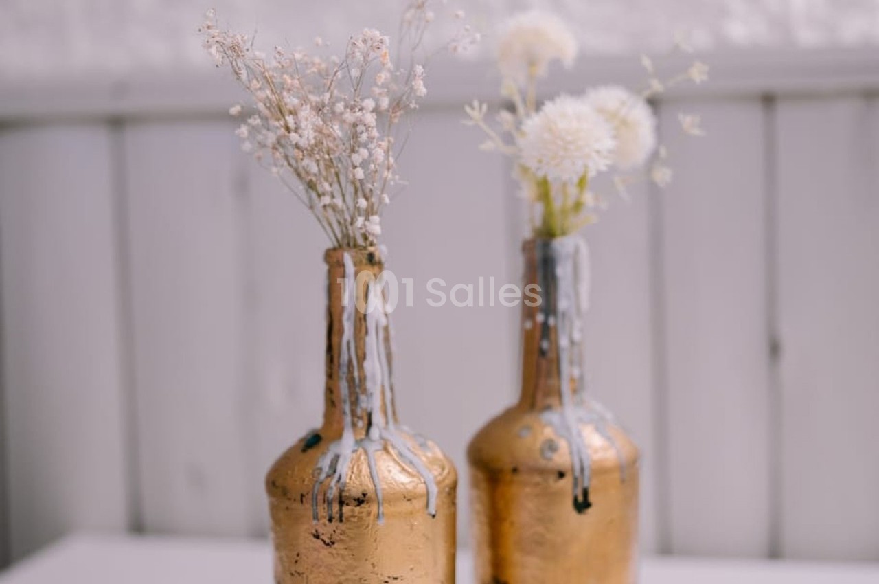 Deux bouteilles dorées recyclées en vases, ornées de cire fondue et contenant des fleurs séchées blanches.