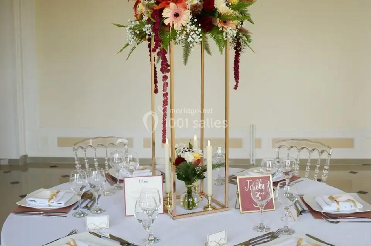Centre de table floral avec structure dorée, bougies et décorations, entouré de vaisselle sur une table ronde dressée.