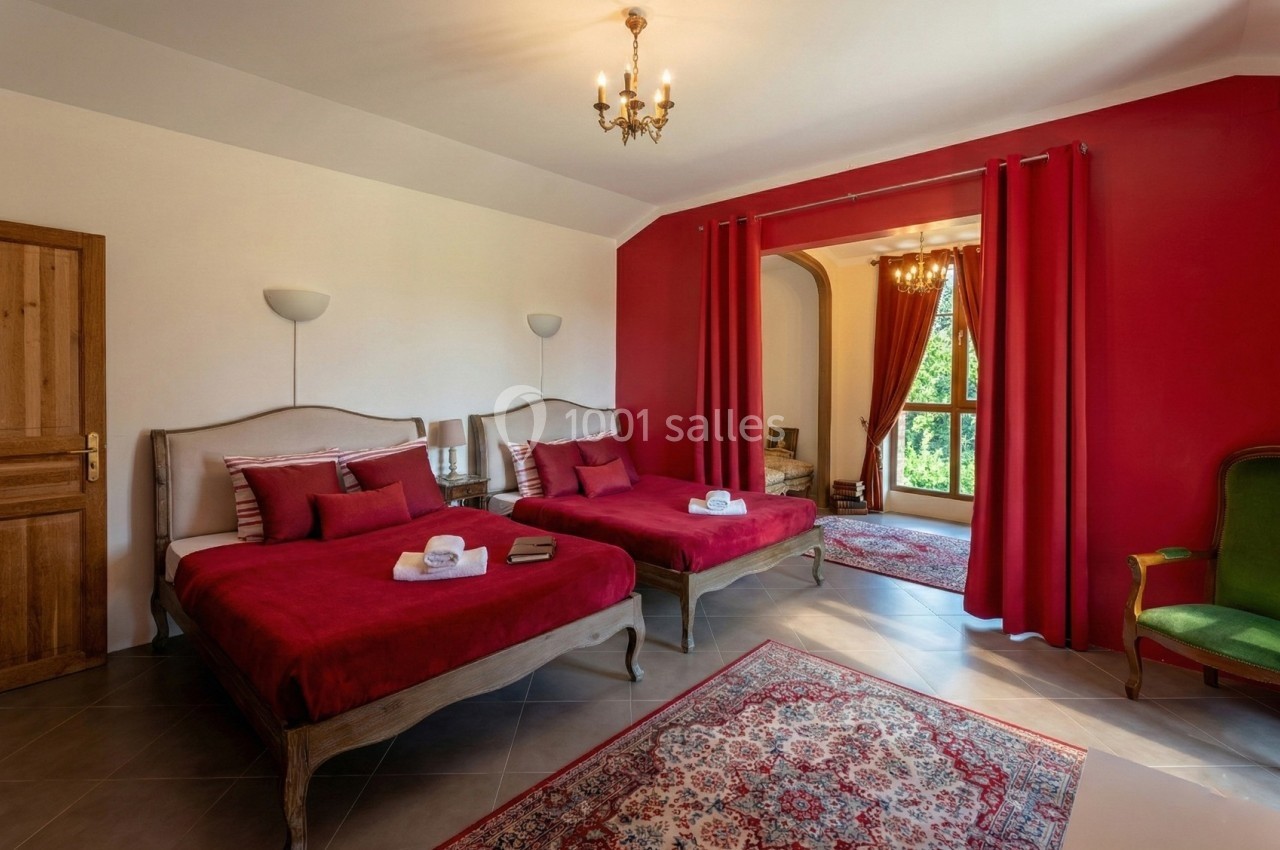 Chambre avec deux lits, murs rouges, fauteuil vert, tapis au sol et grande fenêtre avec rideaux ouverts.