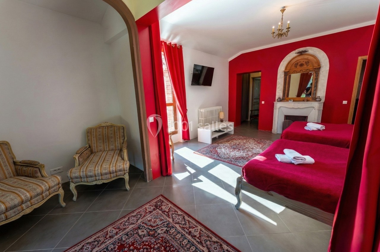 Chambre lumineuse avec murs rouges, deux lits, fauteuils, cheminée, télévision et grande fenêtre avec rideaux rouges.