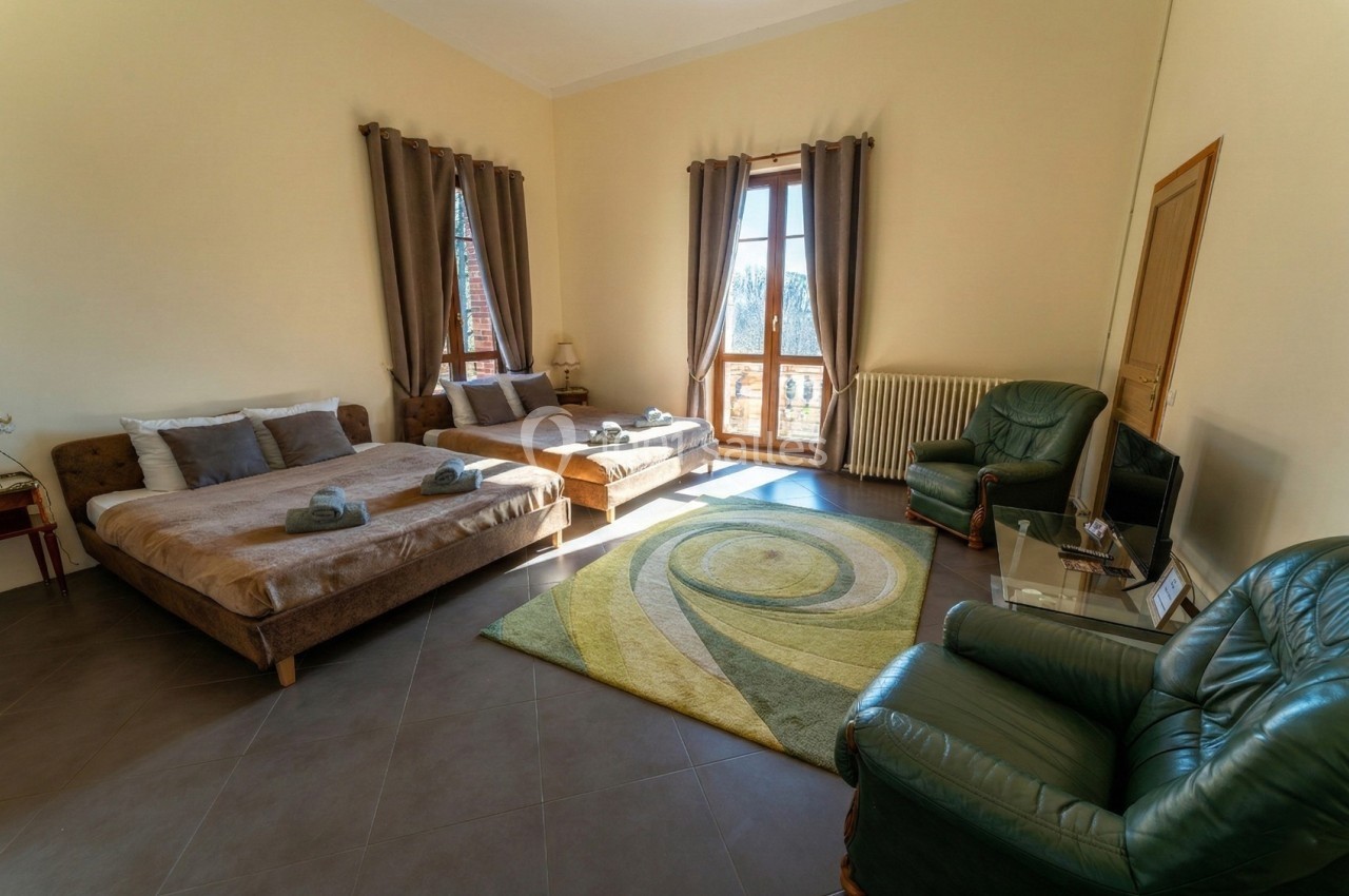 Chambre spacieuse avec deux lits doubles, fauteuils, tapis coloré, grandes fenêtres et rideaux marron.