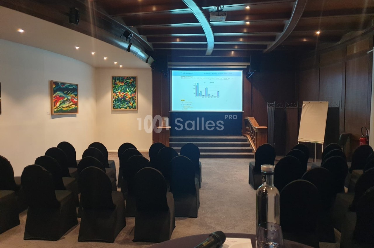 Salle de conférence vide avec des chaises noires, un écran de projection allumé, des tableaux accrochés et un chevalet blanc.