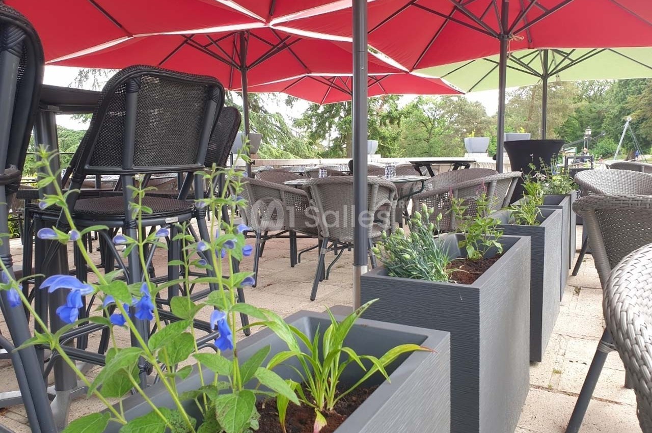 Terrasse aménagée avec tables, chaises en osier, parasols rouges et jardinières contenant des plantes fleuries.