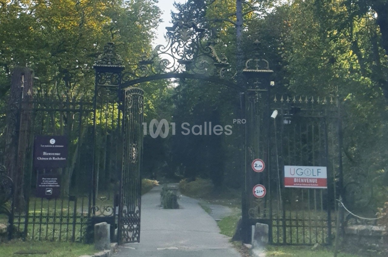 Portail en fer forgé ouvrant sur une allée bordée d'arbres, avec des panneaux d'accueil et de signalisation.