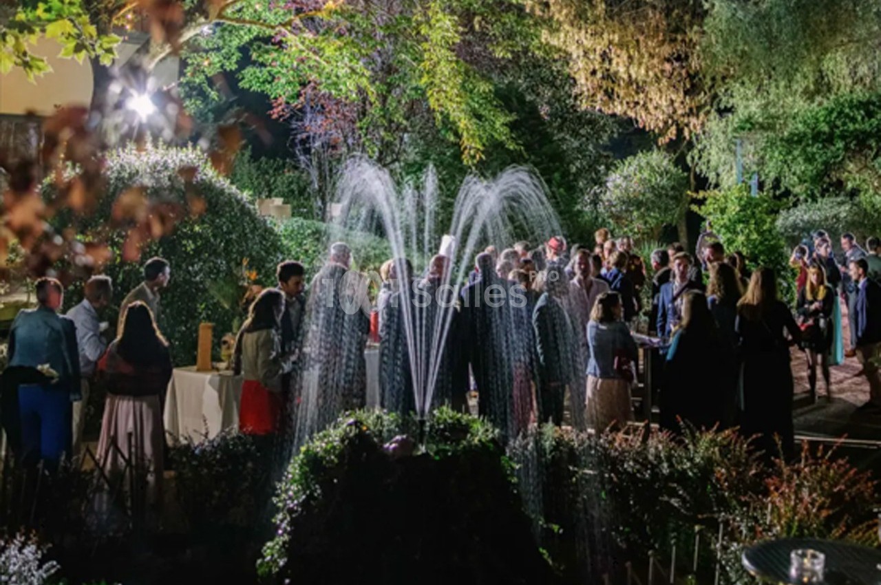 Groupe de personnes rassemblées en soirée dans un jardin éclairé, avec une fontaine illuminée au premier plan.