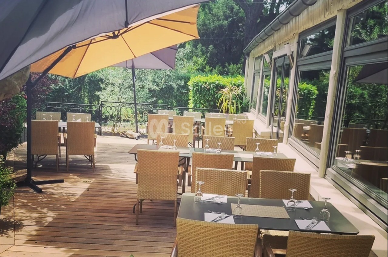 Terrasse en bois avec tables et chaises en osier, ombragée par un grand parasol, entourée de verdure.