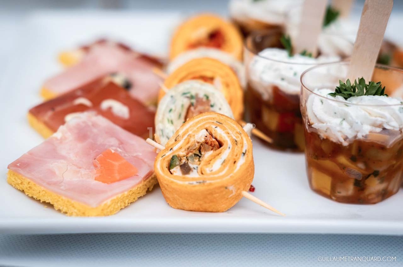 Assortiment de bouchées apéritives comprenant roulés, verrines et toasts sur un plateau blanc.
