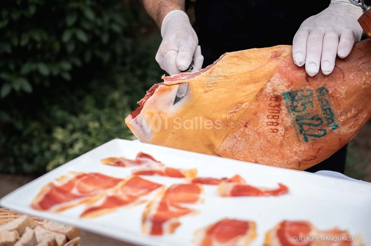 Un cuisinier découpe des tranches fines de jambon cru sur un os, présentées sur une assiette blanche.