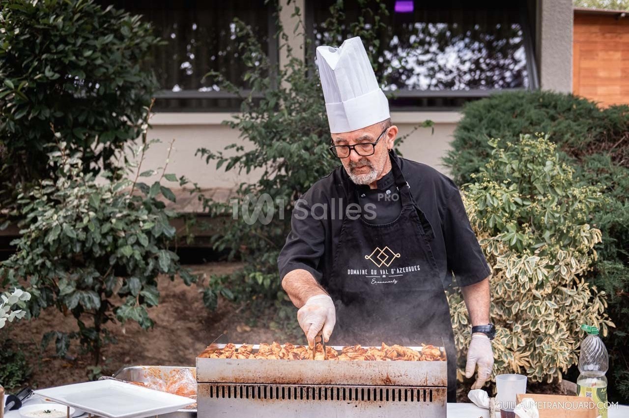 Un chef en tablier et toque cuisine des légumes sur une grande plaque chauffante en extérieur.