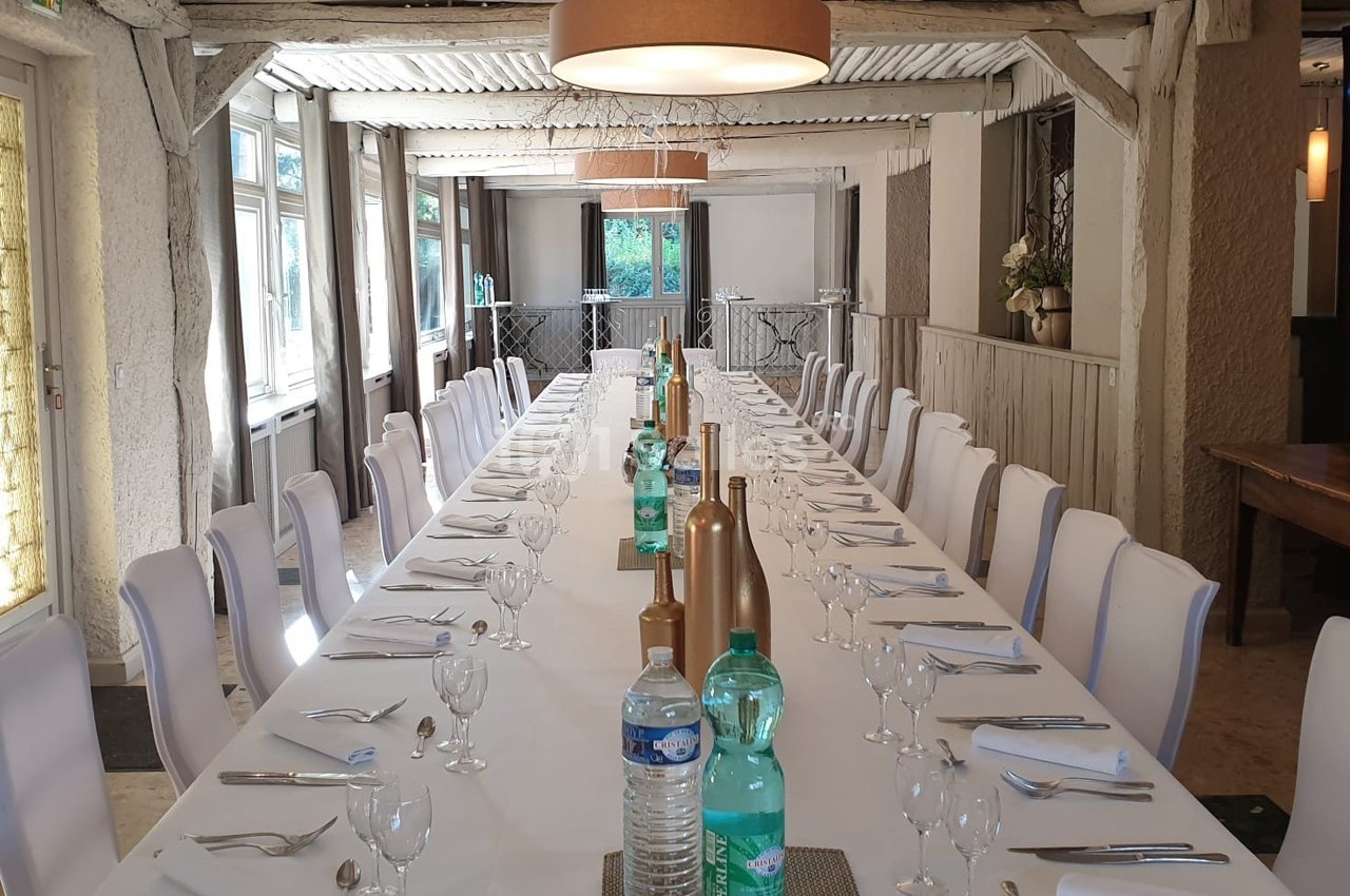 Grande table rectangulaire dressée pour un repas, entourée de chaises blanches, dans une salle lumineuse au style rustique.