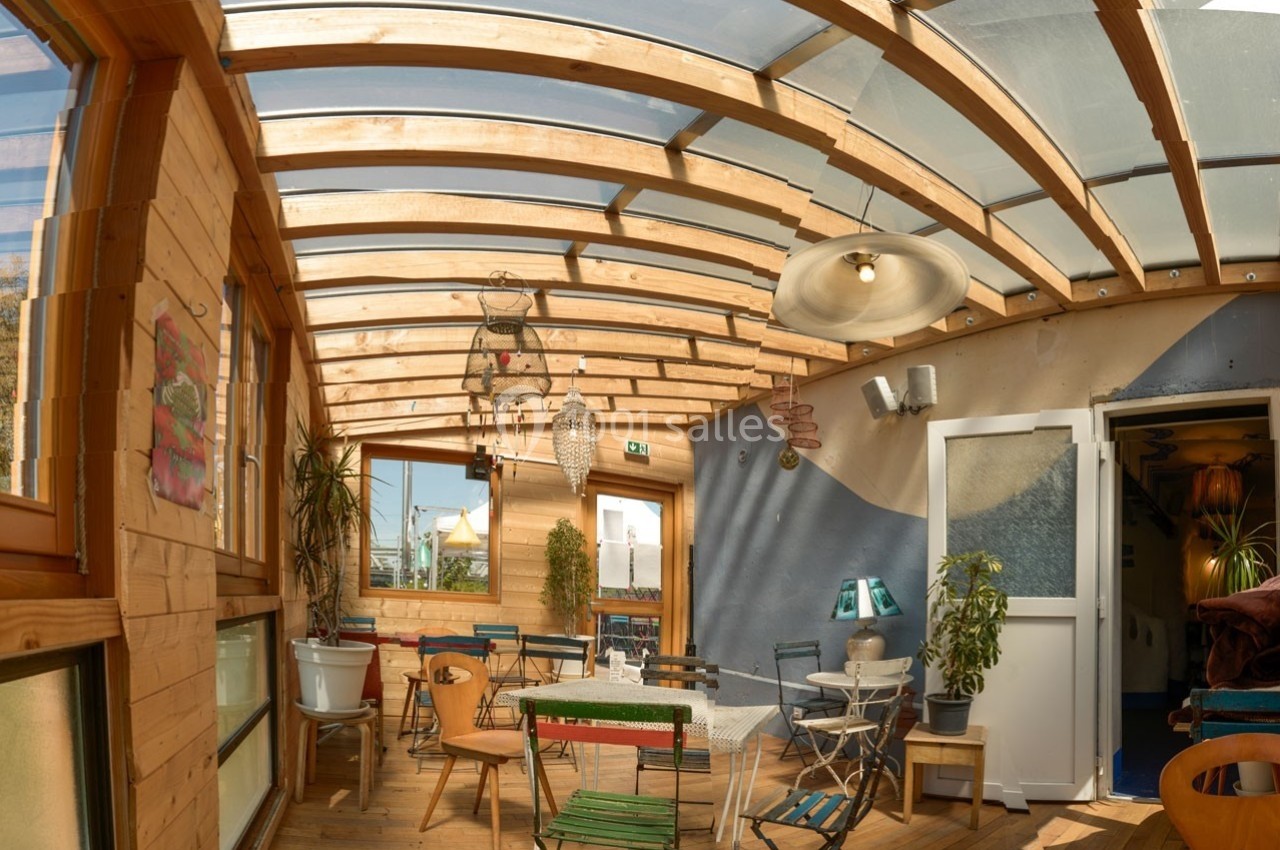 Véranda lumineuse avec toit en verre, mobilier coloré et plantes, donnant sur une porte ouverte vers l'intérieur.