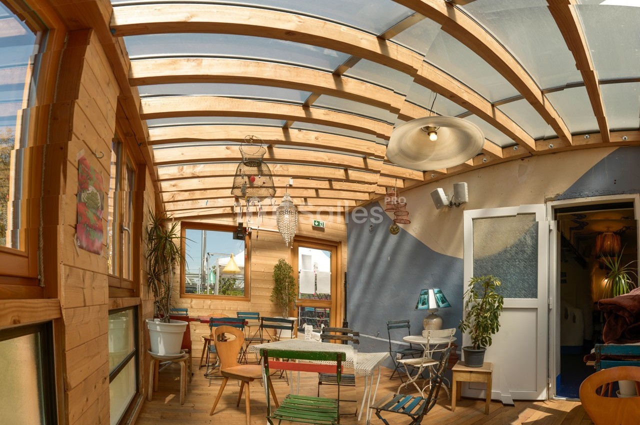 Véranda lumineuse avec toit en verre, mobilier coloré et plantes, donnant sur une porte ouverte vers l'intérieur.