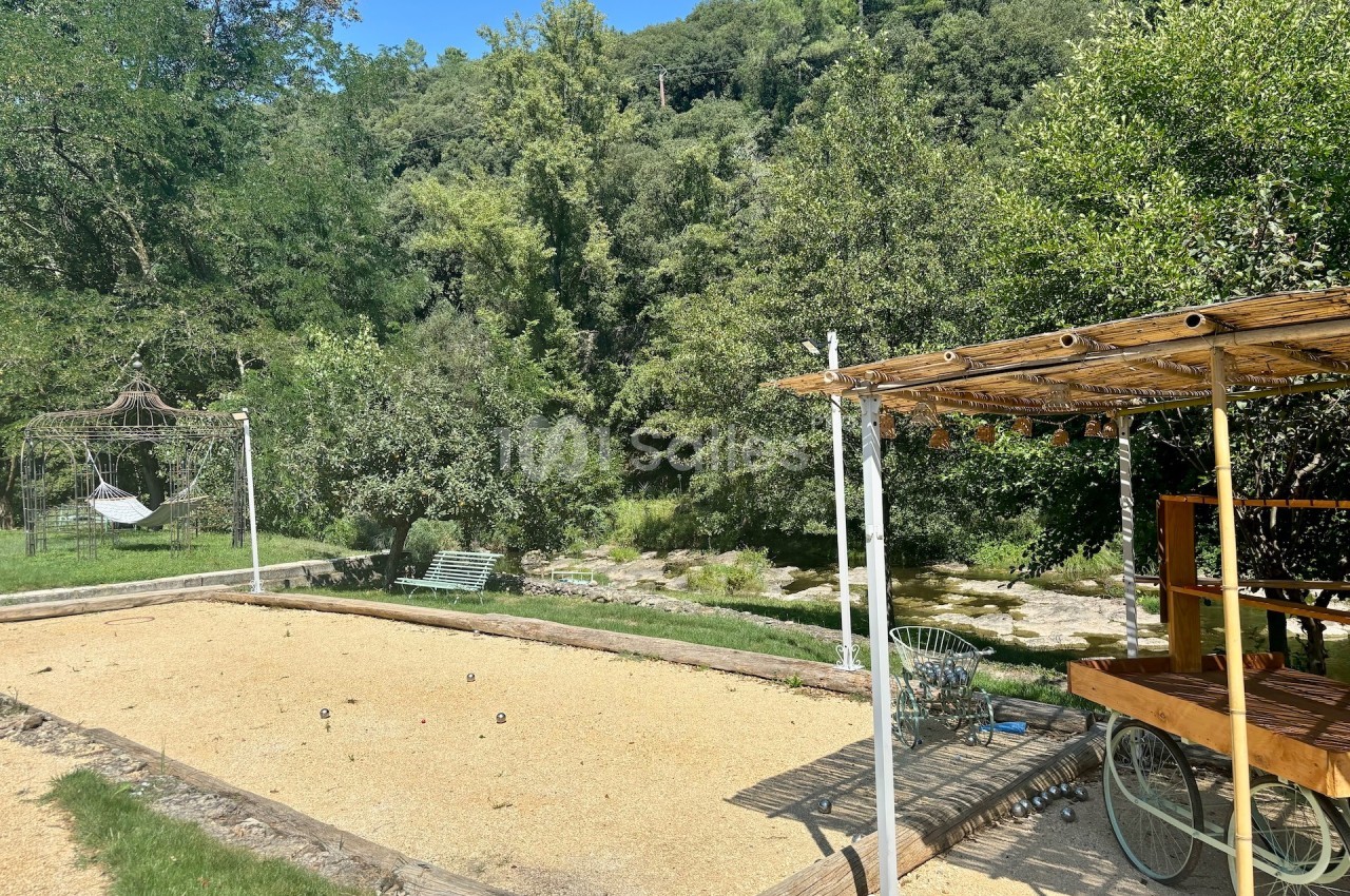 Terrain de pétanque ensoleillé avec pergola en bois, entouré de verdure et d'arbres, près d'une rivière.