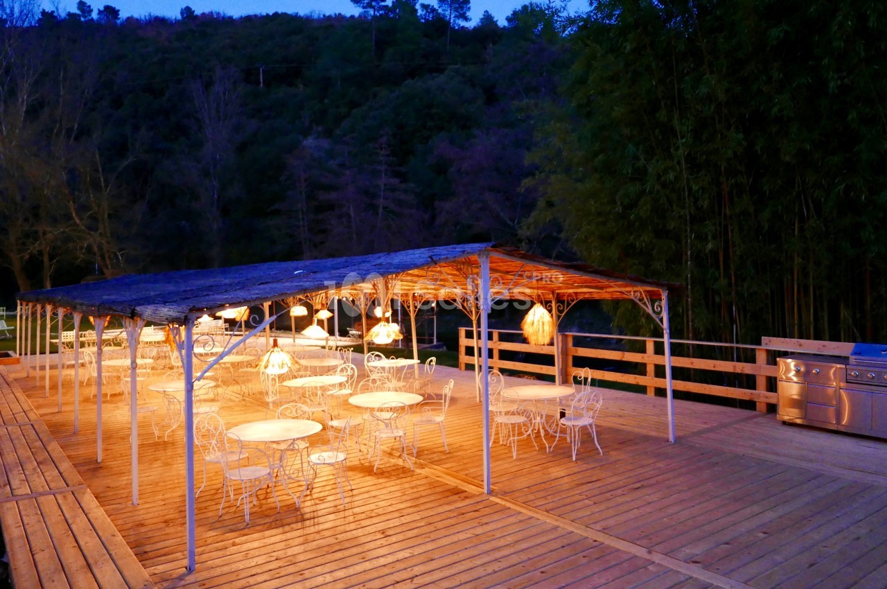 Terrasse en bois éclairée le soir, avec des tables et chaises blanches sous une pergola, entourée de végétation.