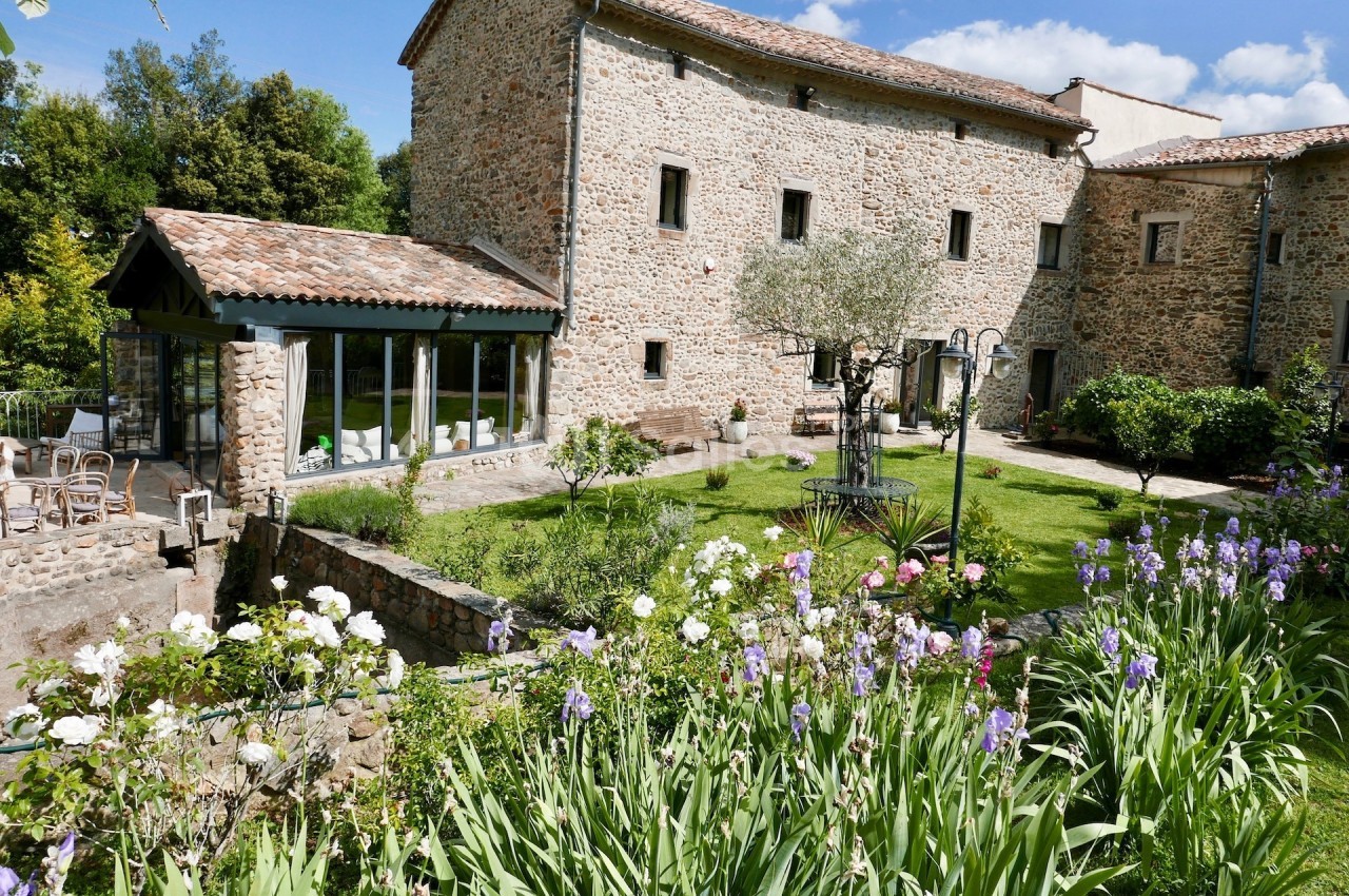 Jardin fleuri avec pelouse, arbres et terrasse, entouré d'un bâtiment en pierre sous un ciel dégagé.