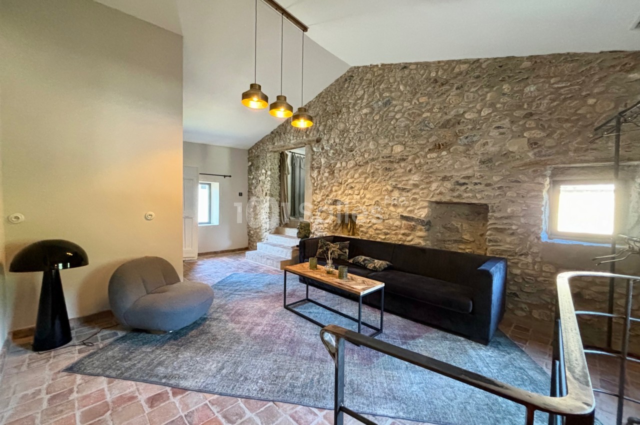 Salon avec murs en pierre, canapé noir, fauteuil gris, tapis bleu, éclairage suspendu et sol en carreaux de terre cuite.