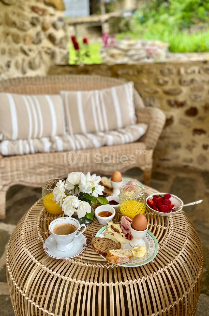 Petit-déjeuner composé de pain, charcuterie, œufs, fruits et café, servi sur une table en rotin dans un cadre champêtre.