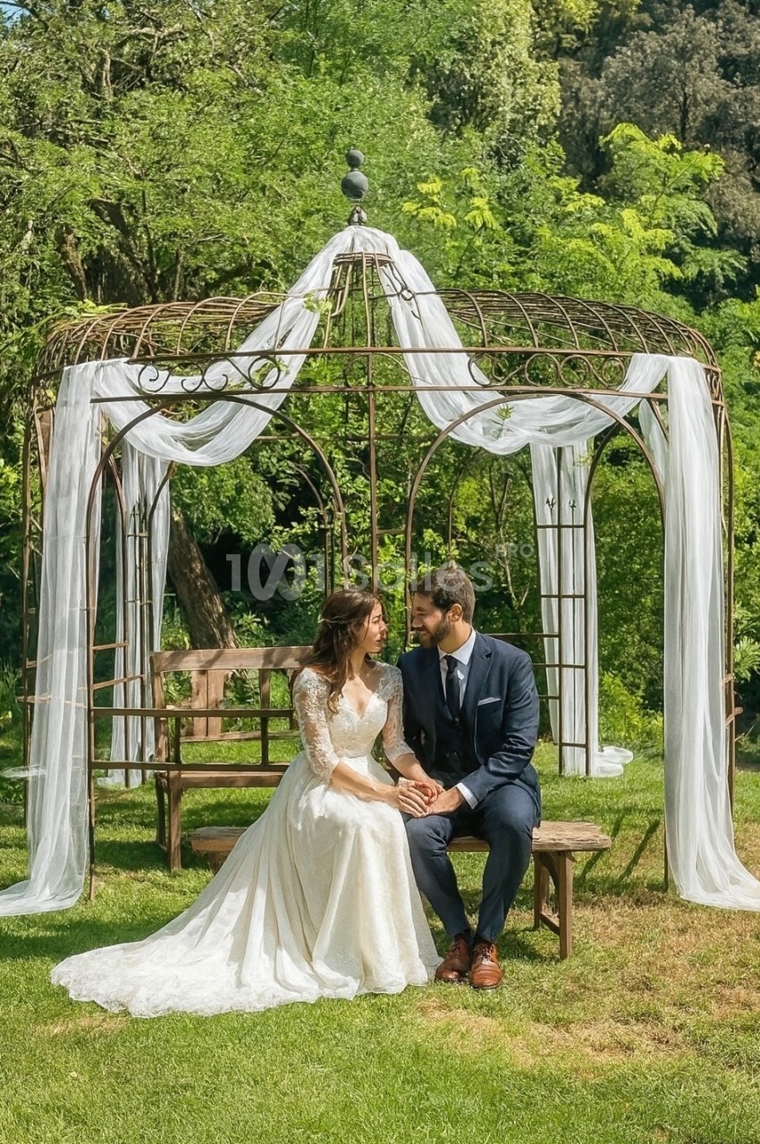 Un couple assis sous une tonnelle décorée de voilages blancs dans un jardin verdoyant.