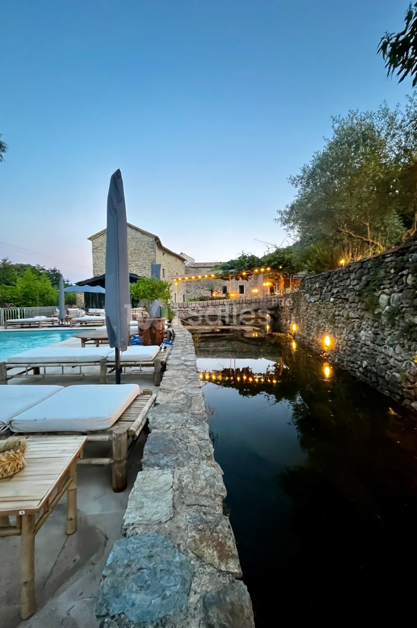 Terrasse avec piscine, chaises longues, parasol et bassin d'eau entouré de pierres, éclairée à la tombée de la nuit.