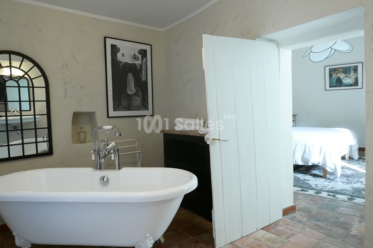 Salle de bain avec baignoire sur pieds, miroir en arche, et porte ouverte donnant sur une chambre lumineuse.