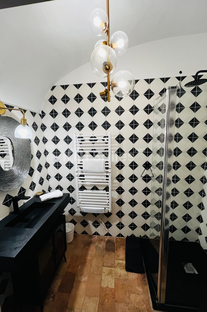 Salle de bain moderne avec carrelage géométrique noir et blanc, douche vitrée, lavabo noir et miroir rond texturé.