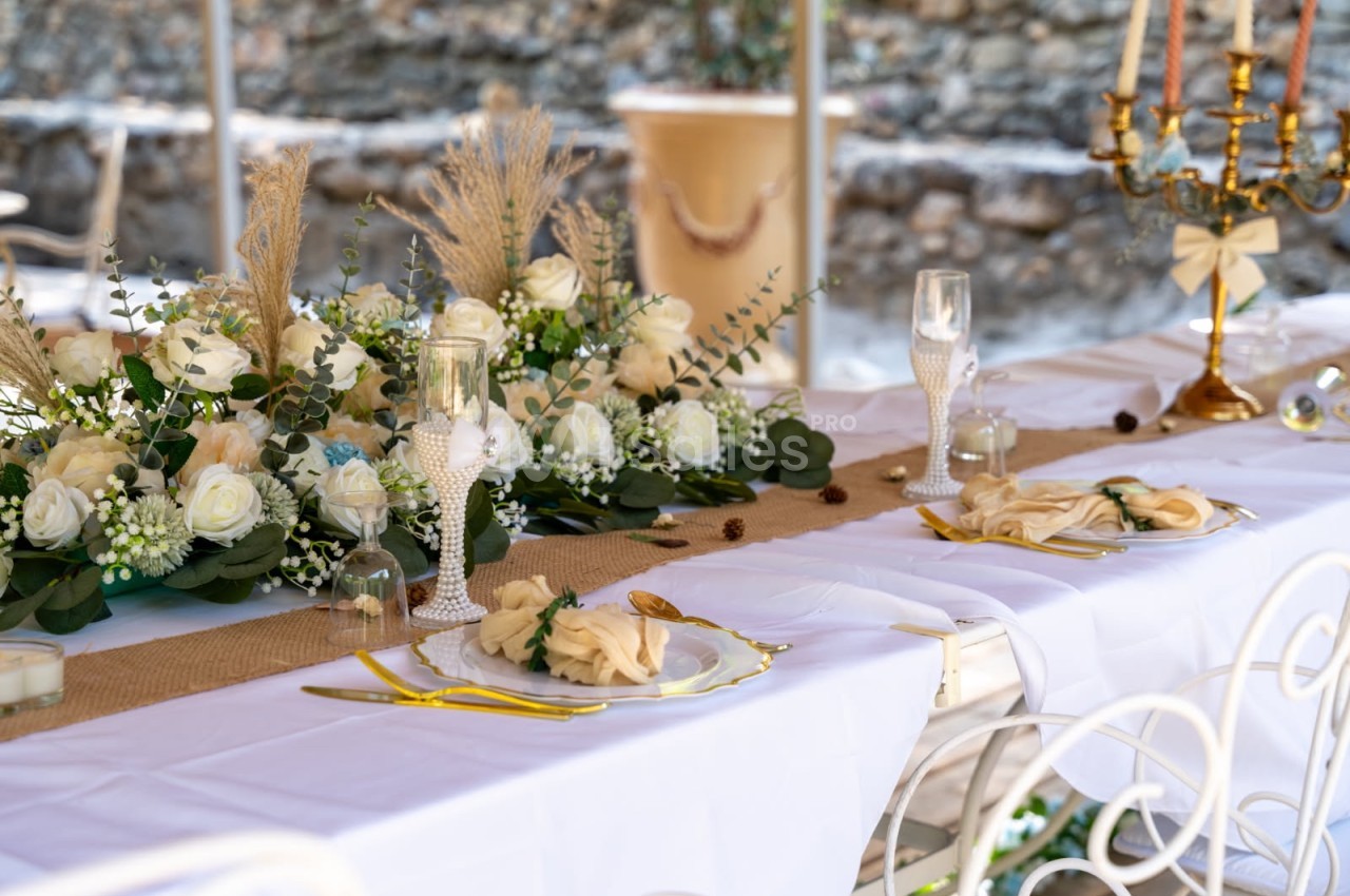 Table décorée pour un événement, avec nappes blanches, fleurs, bougies et couverts dorés dans un cadre extérieur.