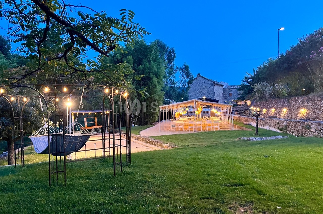 Jardin éclairé au crépuscule avec un hamac suspendu, une pergola lumineuse et des tables dressées en arrière-plan.