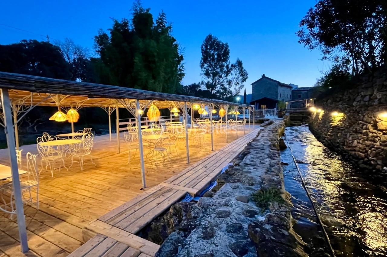 Terrasse en bois éclairée par des lampes suspendues, bordée d'un ruisseau et entourée de végétation au crépuscule.