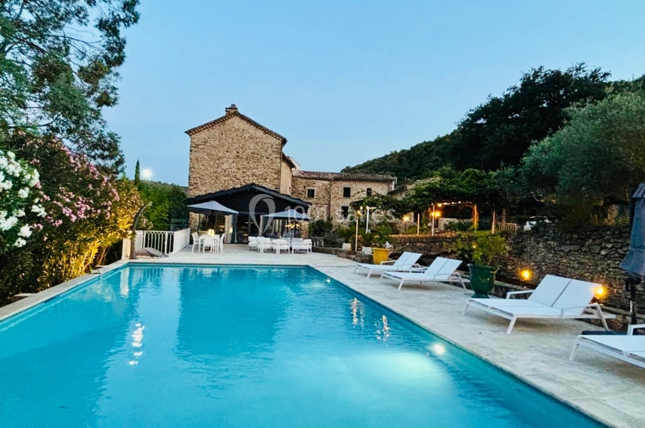 Piscine extérieure entourée de chaises longues et végétation, avec une maison en pierre en arrière-plan au crépuscule.