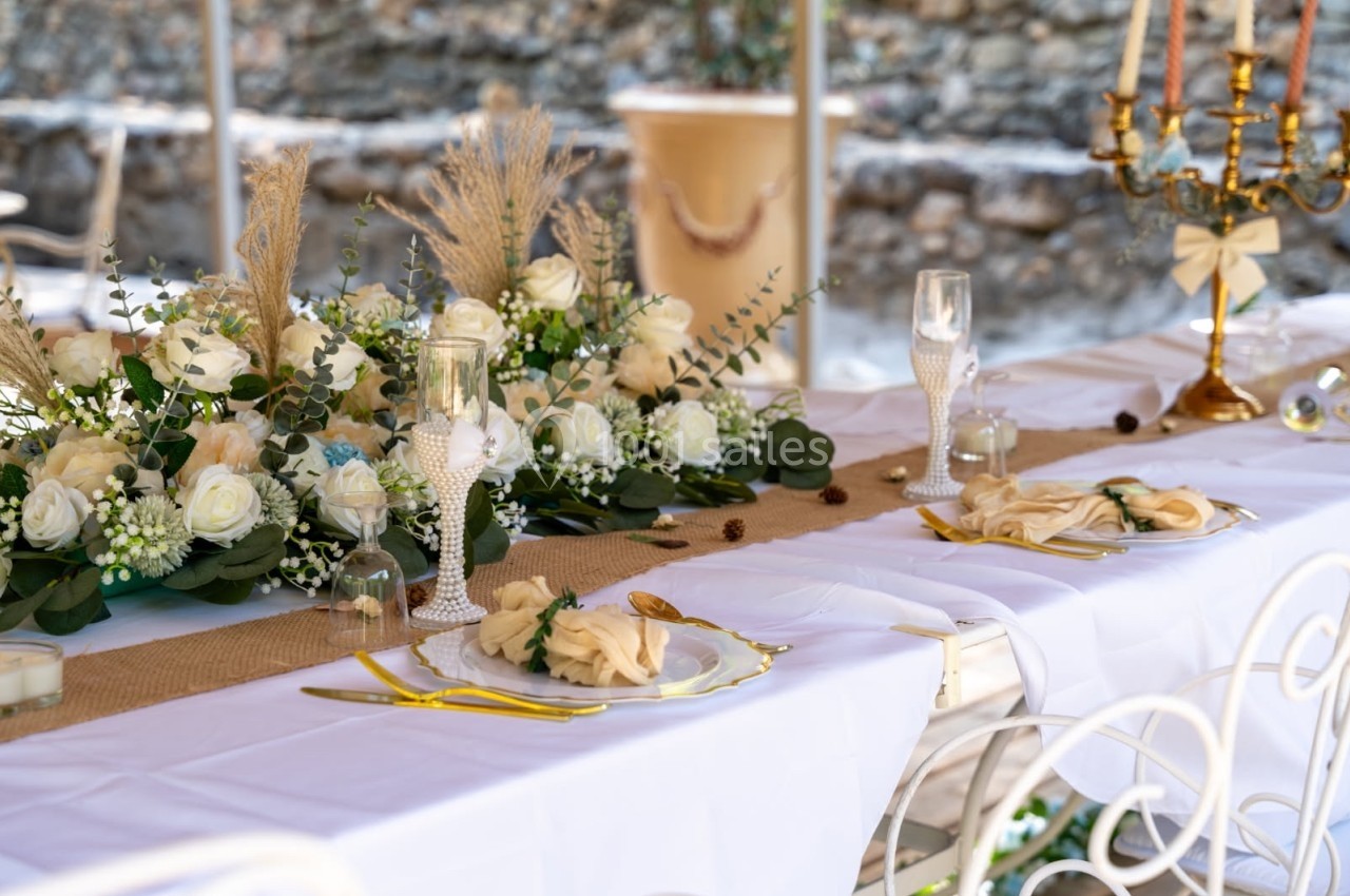 Table décorée pour un événement, avec nappes blanches, fleurs blanches, couverts dorés et chandeliers en arrière-plan.