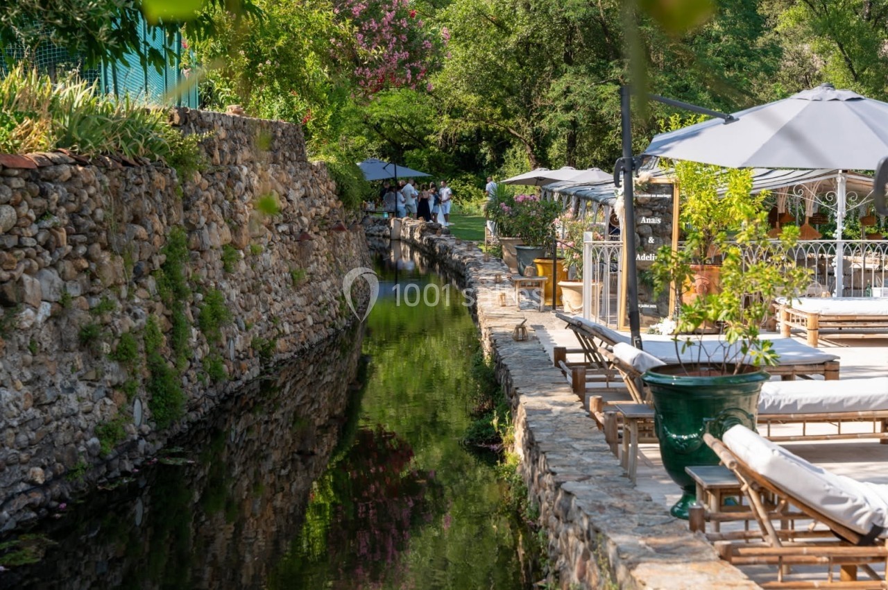 Canal bordé de pierres avec des chaises longues et parasols, entouré de verdure et d'arbres en arrière-plan.