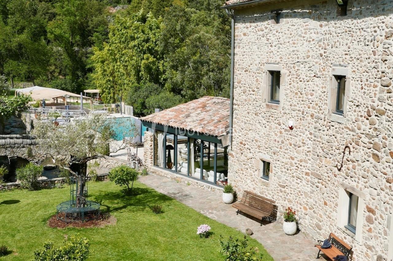 Cour intérieure d'une maison en pierre avec jardin verdoyant, arbres fruitiers, bancs et piscine en arrière-plan.