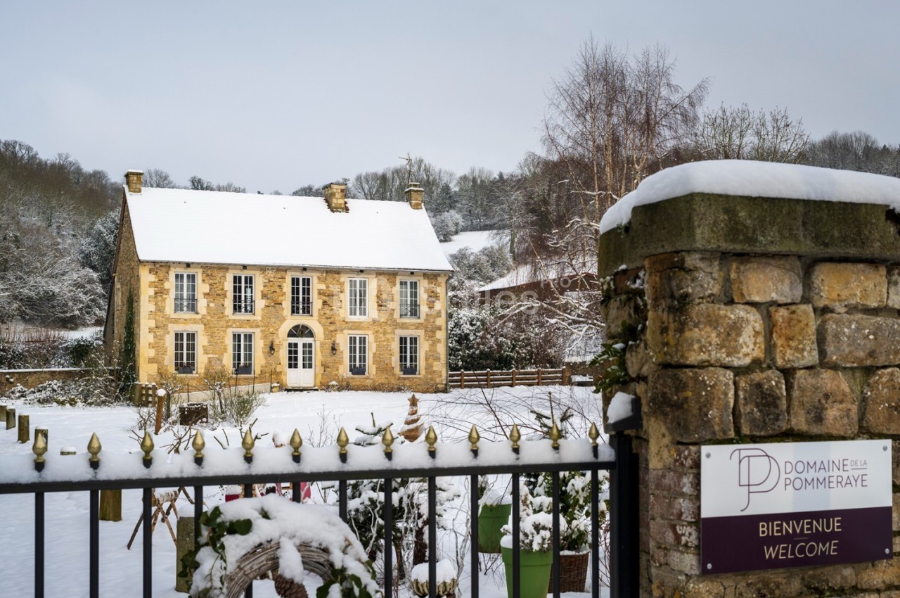 Grande maison en pierre entourée de neige, vue derrière un portail avec un panneau indiquant ’Domaine de la Pommeraye’.