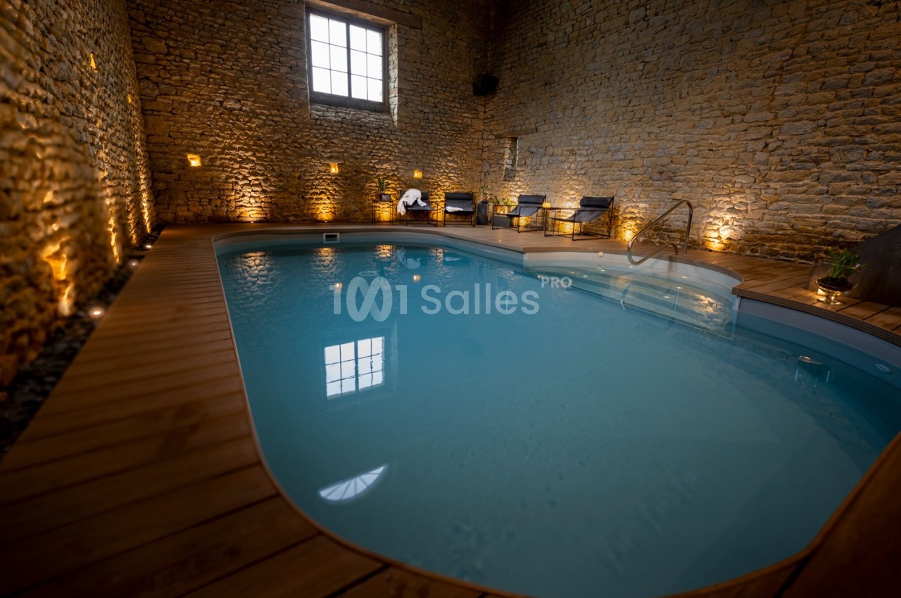 Piscine intérieure éclairée dans une pièce aux murs en pierre avec un sol en bois et des transats disposés autour.