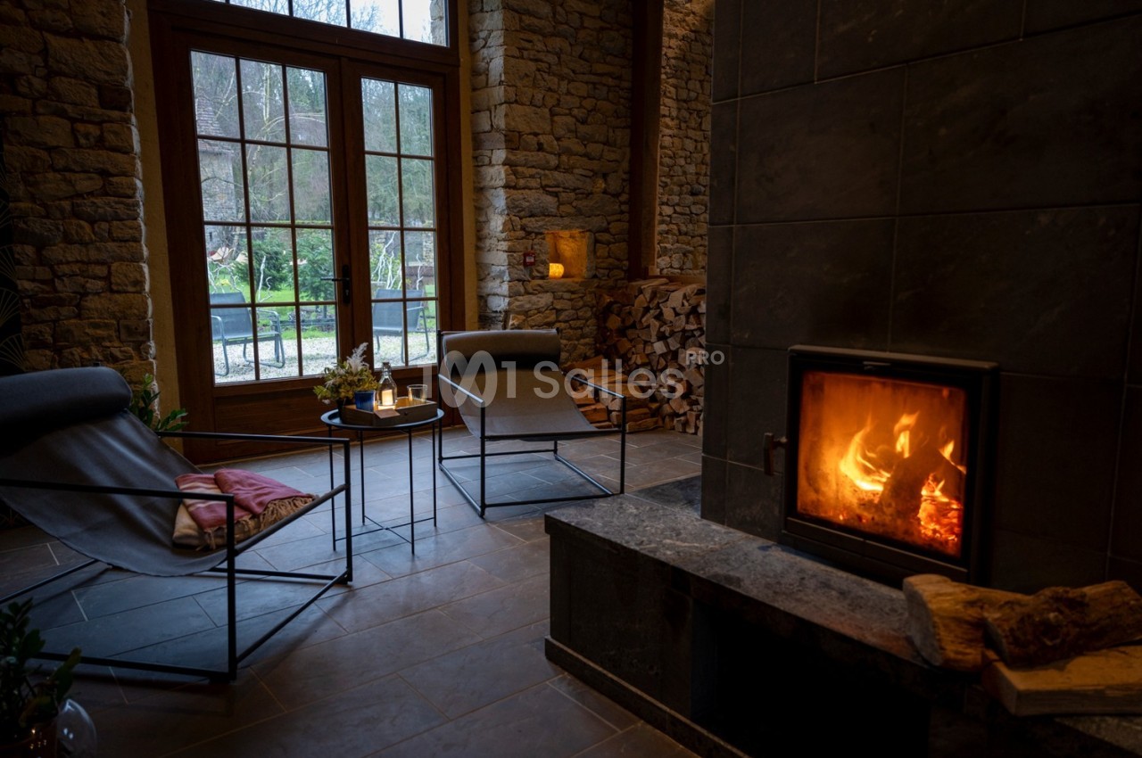 Salon chaleureux avec poêle à bois allumé, fauteuils modernes, murs en pierre et vue sur un jardin à travers de grandes…