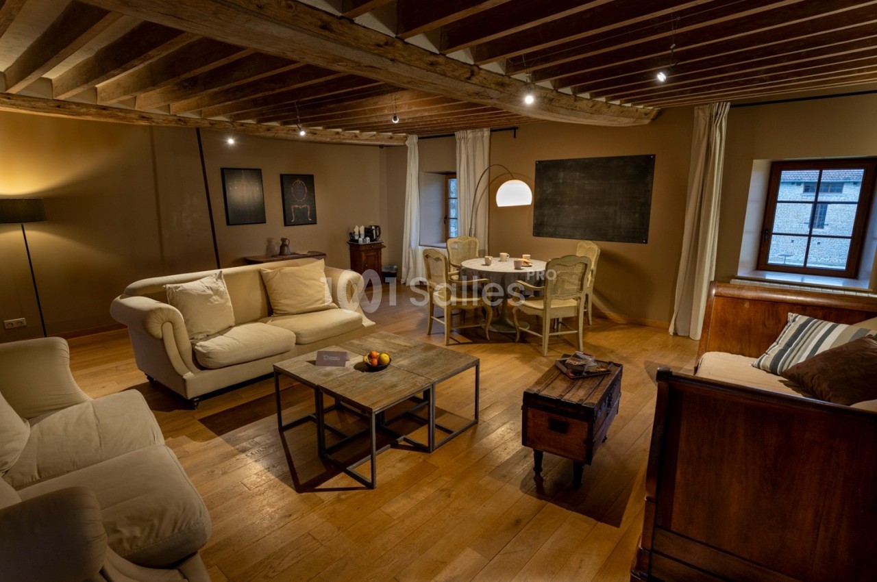 Salon chaleureux avec poutres apparentes, mobilier en bois, canapés clairs et table à manger près d'une fenêtre.