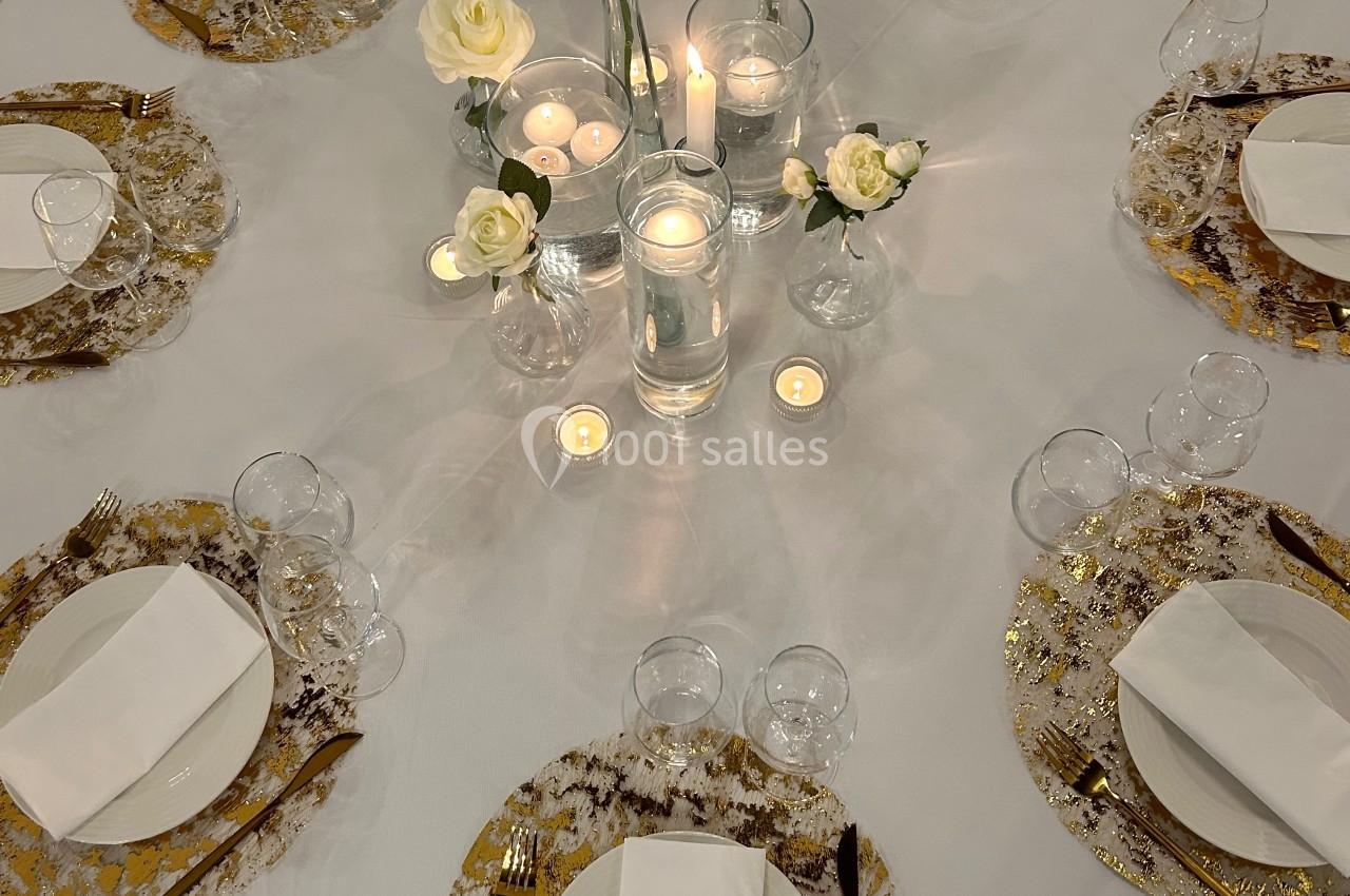 Table ronde élégamment dressée avec des assiettes dorées, des couverts dorés, des roses blanches et des bougies allumées.