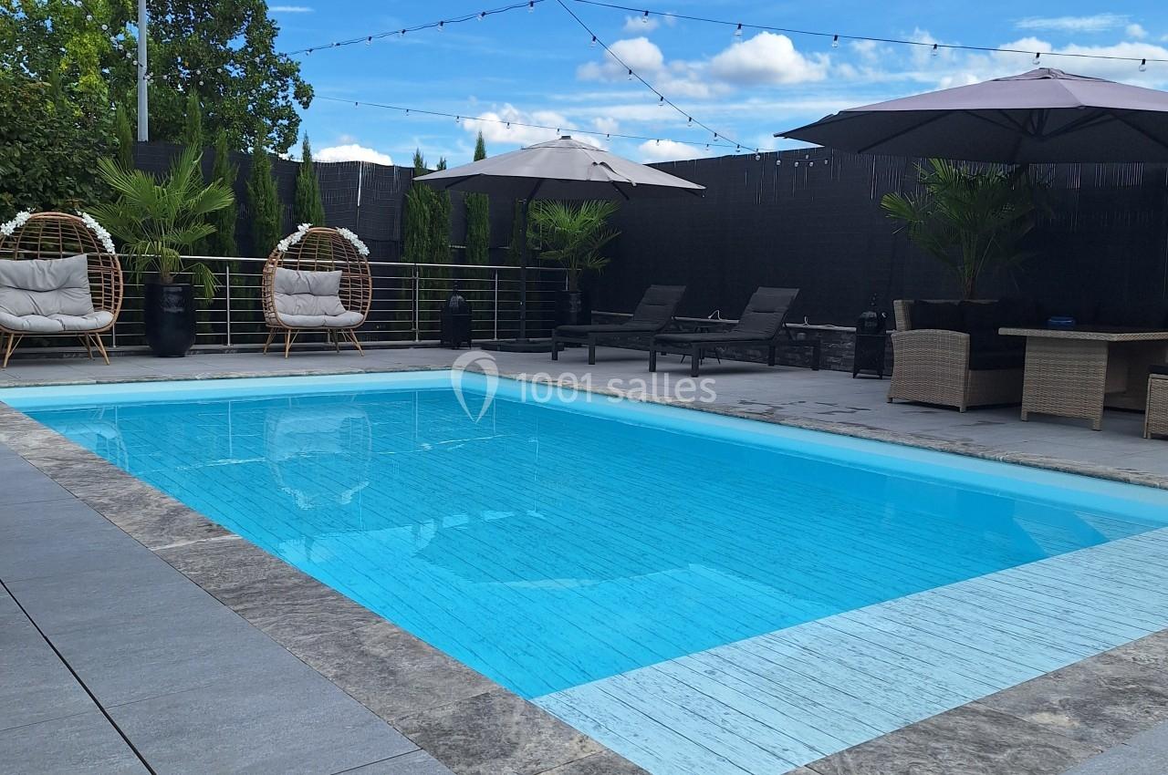 Piscine extérieure entourée de carrelage gris, avec parasols, fauteuils suspendus et ciel bleu en arrière-plan.