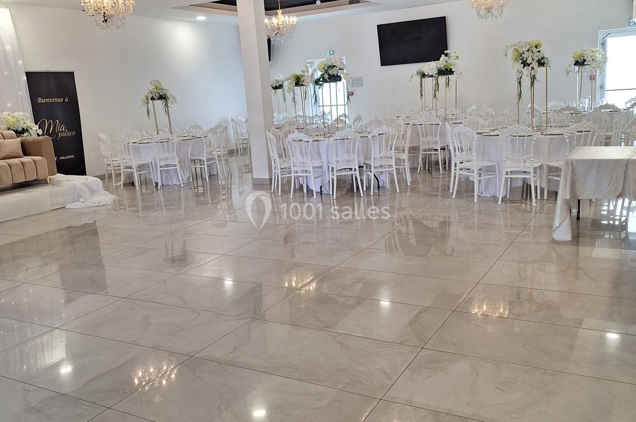 Salle de réception lumineuse avec tables et chaises blanches, décorations florales et lustres suspendus.