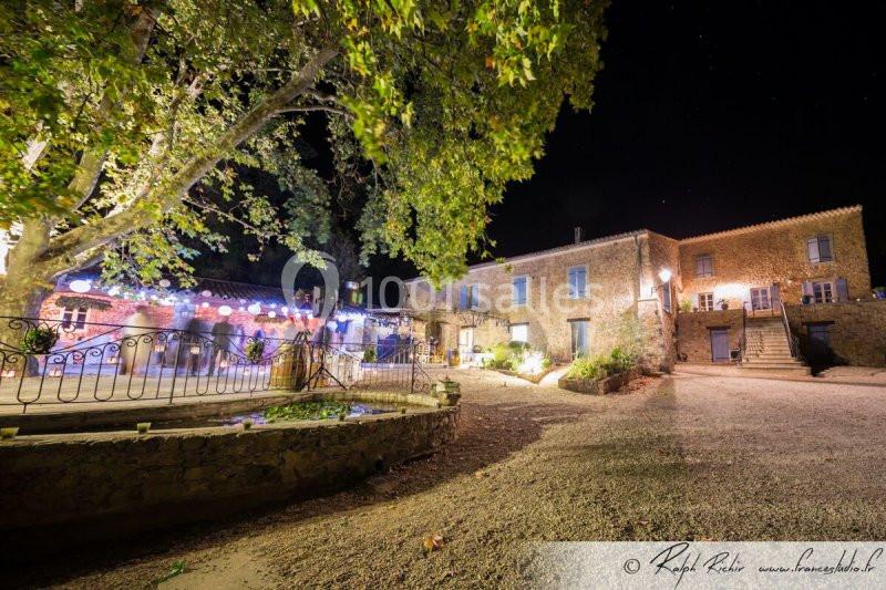 Cour d'une maison en pierre éclairée la nuit, avec un grand arbre, des guirlandes lumineuses et un sol en gravier.