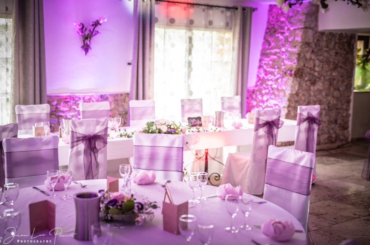 Salle de réception décorée pour un événement, avec des tables dressées et des chaises ornées de nœuds violets.