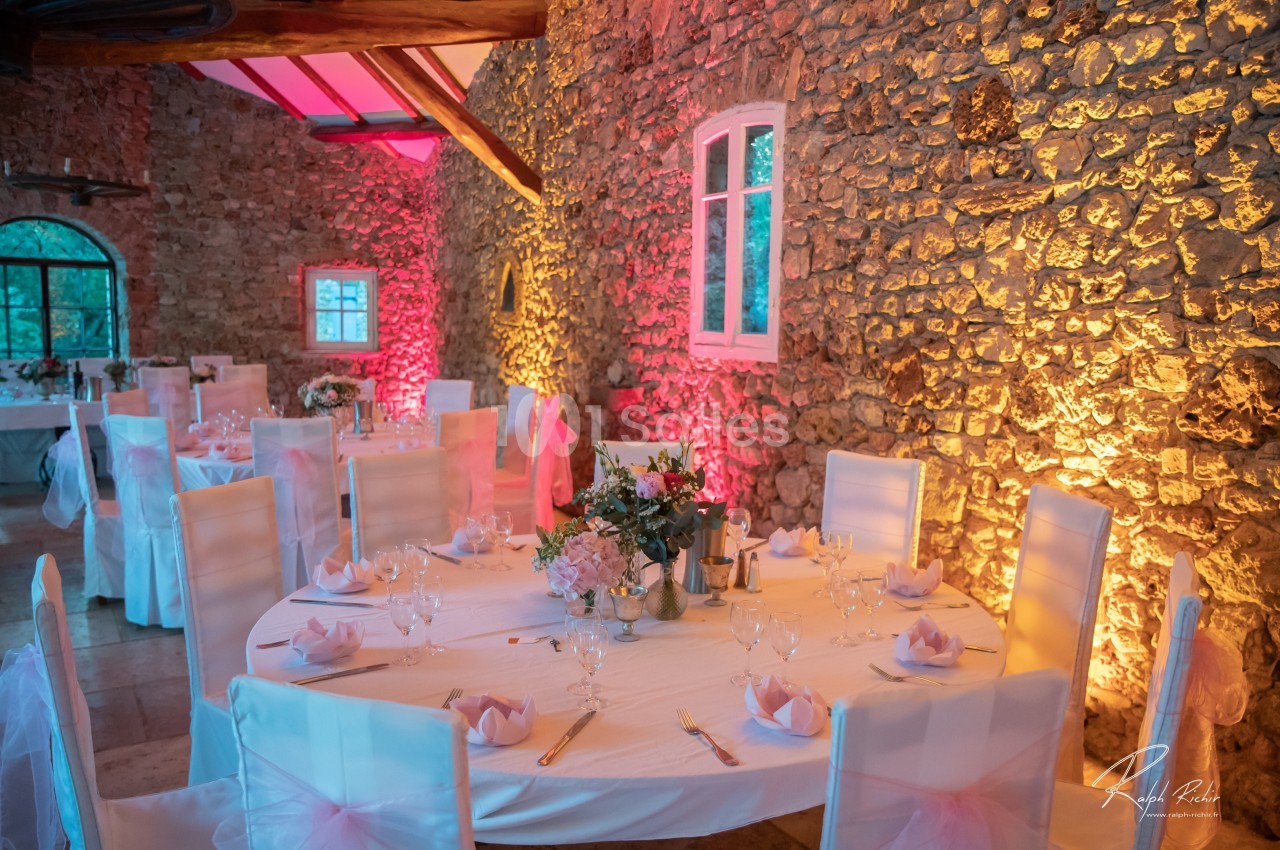 Salle de réception en pierre avec tables rondes décorées de nappes blanches, fleurs et éclairage coloré.