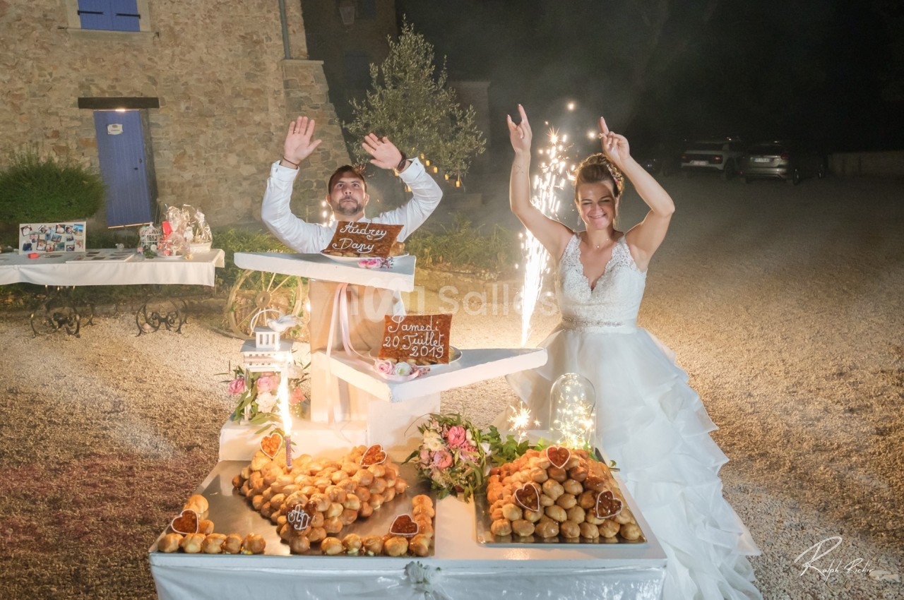 Un couple de mariés souriants célèbre devant une table garnie de desserts et de décorations lumineuses.
