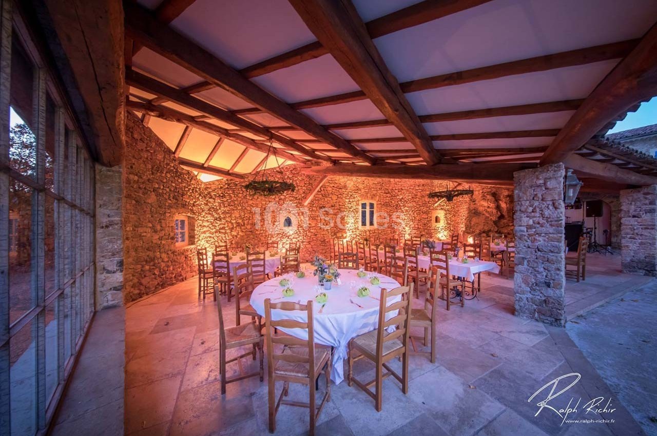 Salle en pierre avec tables rondes dressées, éclairage chaleureux et plafond en poutres apparentes.