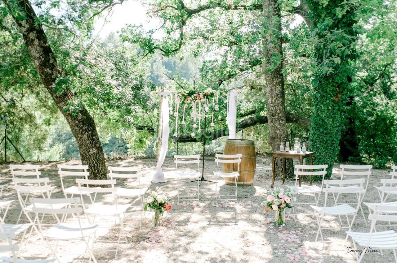 Chaises blanches disposées devant une arche décorée de fleurs et de voilages, dans un cadre boisé en plein air.