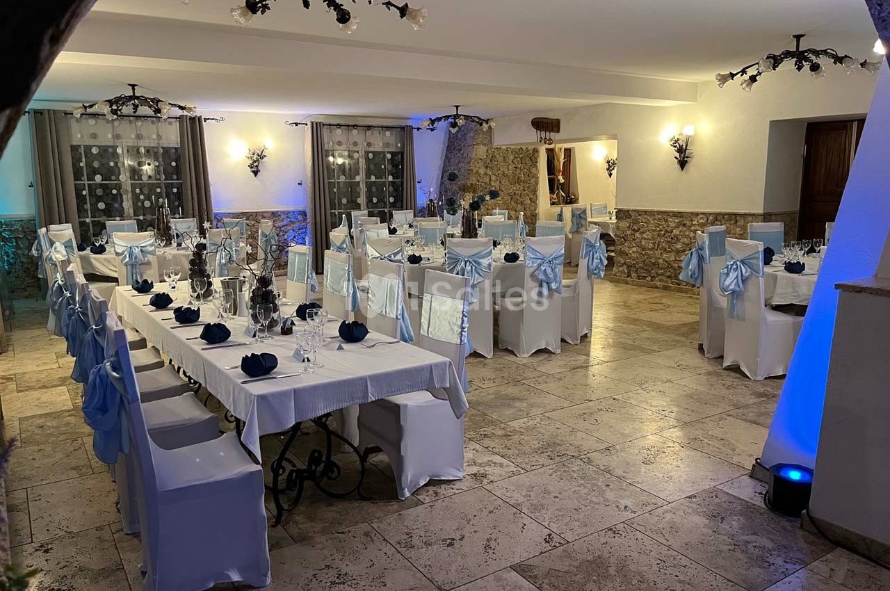 Salle de réception décorée avec des nappes blanches et des chaises ornées de nœuds bleus, éclairage chaleureux.