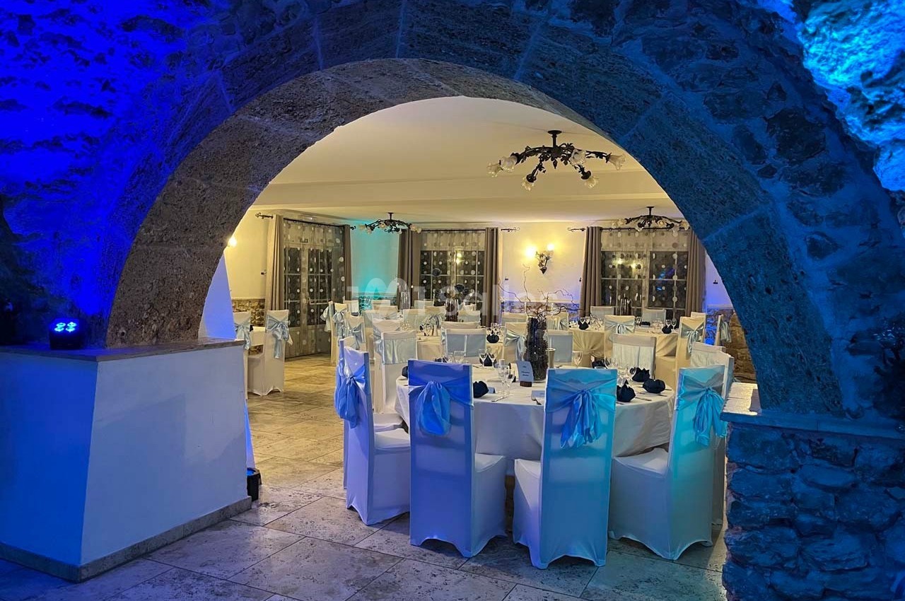 Salle de réception élégamment décorée, avec des tables dressées et des chaises blanches sous un éclairage bleu tamisé.