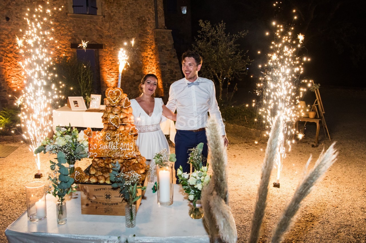 Un couple souriant devant une pièce montée éclairée par des bougies et des feux d'artifice en extérieur, de nuit.