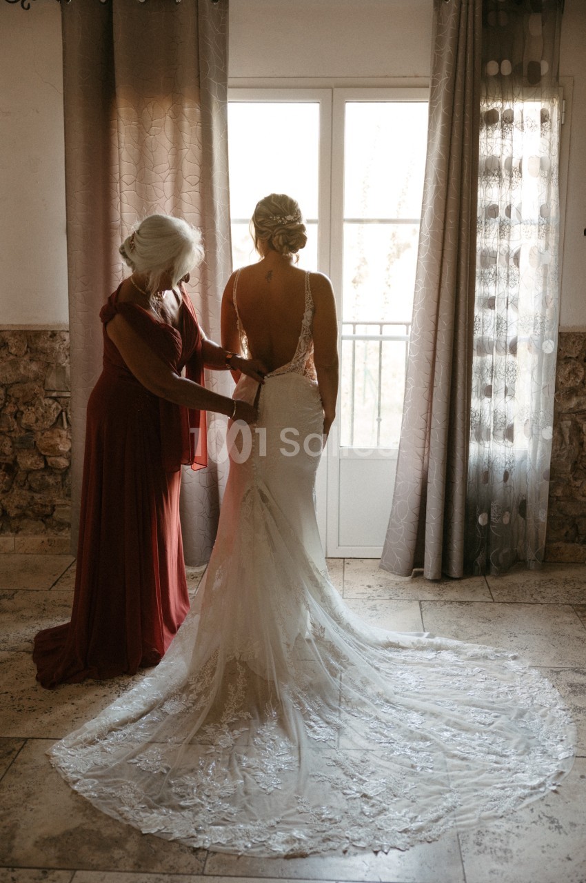 Une femme aide une mariée à ajuster sa robe dans une pièce lumineuse avec des rideaux et une porte-fenêtre en arrière-plan.
